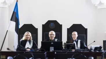 Kolm kohtunikku istung kohtusaalis laua taga. Laua kõrval seisab Eesti lipp. 